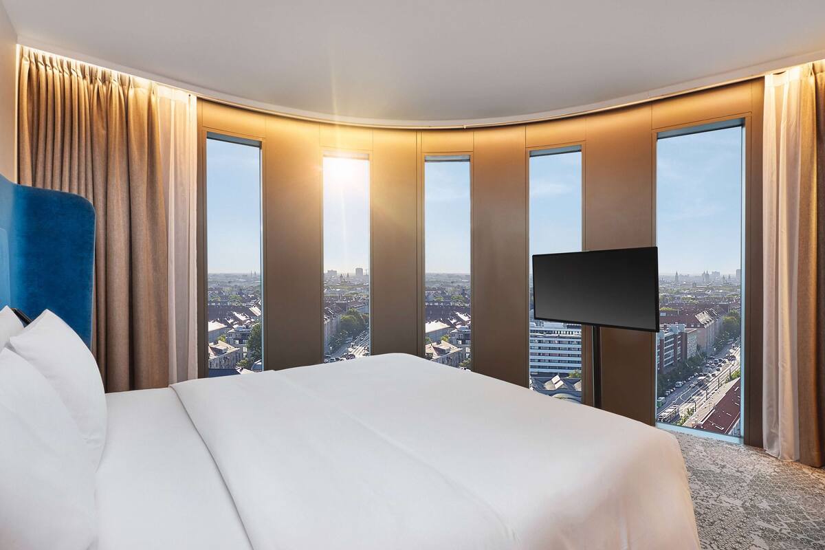 Habitación de hotel con una cama grande, un televisor de pantalla plana y vistas a la ciudad a través de amplias ventanas.