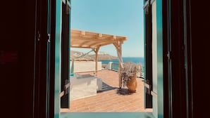 Ett öppet fönster med utsikt över en träpergola, en krukväxt och en terrass med kakelplattor, med utsikt över havet.