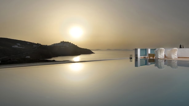 En vacker utsikt över vattnet med en klar himmel, ett stilla vatten som speglar solen och en modern byggnad med en pool och ett område med sittplatser.
