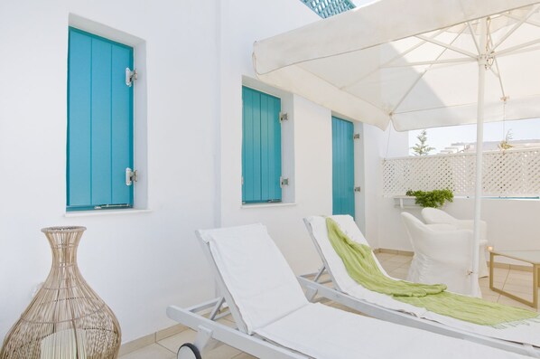 A sunlit outdoor lounge area with two white lounge chairs, a wicker vase, and a white umbrella providing shade.