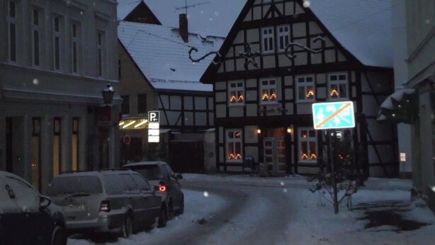 Un edificio tradicional de entramado de madera con techo cubierto de nieve, coches aparcados y un cartel de estacionamiento visible.