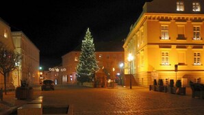 Una plaza adoquinada con un gran árbol de Navidad, edificios con ventanas iluminadas y un coche estacionado en la calle.