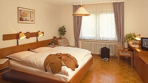 A hotel room with a bed, bedside lamps, a desk with a television, a chair, a radiator, and a window with curtains. - Hörster Krug (Lage)