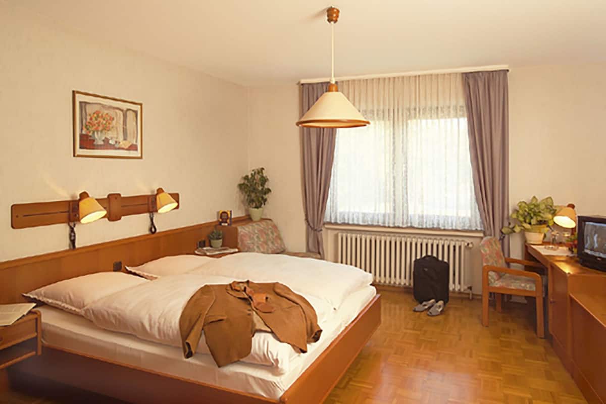A hotel room with a bed, bedside lamps, a desk with a television, a chair, a radiator, and a window with curtains.