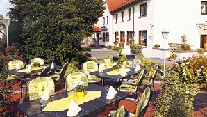 An outdoor seating area with tables and chairs, surrounded by greenery and a building with a red-tiled roof. - Hörster Krug (Lage)
