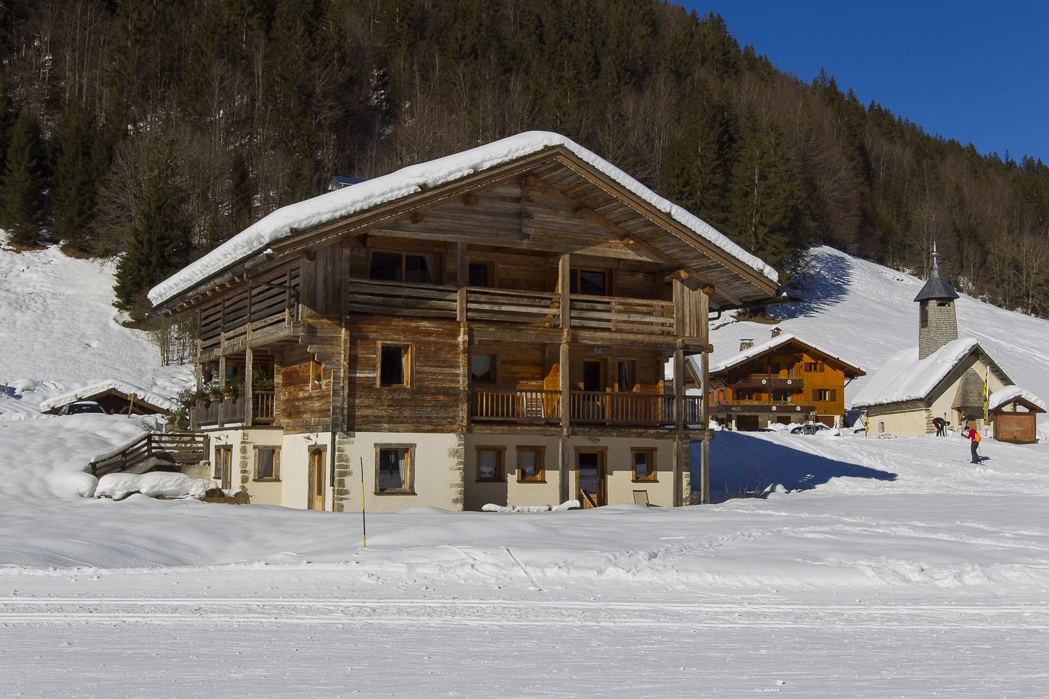 A wooden chalet with a balcony, surrounded by snow-covered terrain and a forested mountain backdrop.