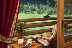 Una terraza de madera con una silla, una mesa con un hervidor y un plato de comida, y vistas a un paisaje verde con árboles y un edificio pequeño.