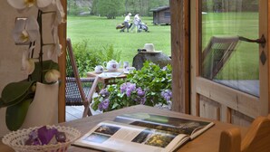 A room with a view of a grassy yard, a wooden table with a book, a vase with flowers, and a window with a view of people in the distance. - Les Fermes de Pierre et Anna (Le Grand-Bornand)