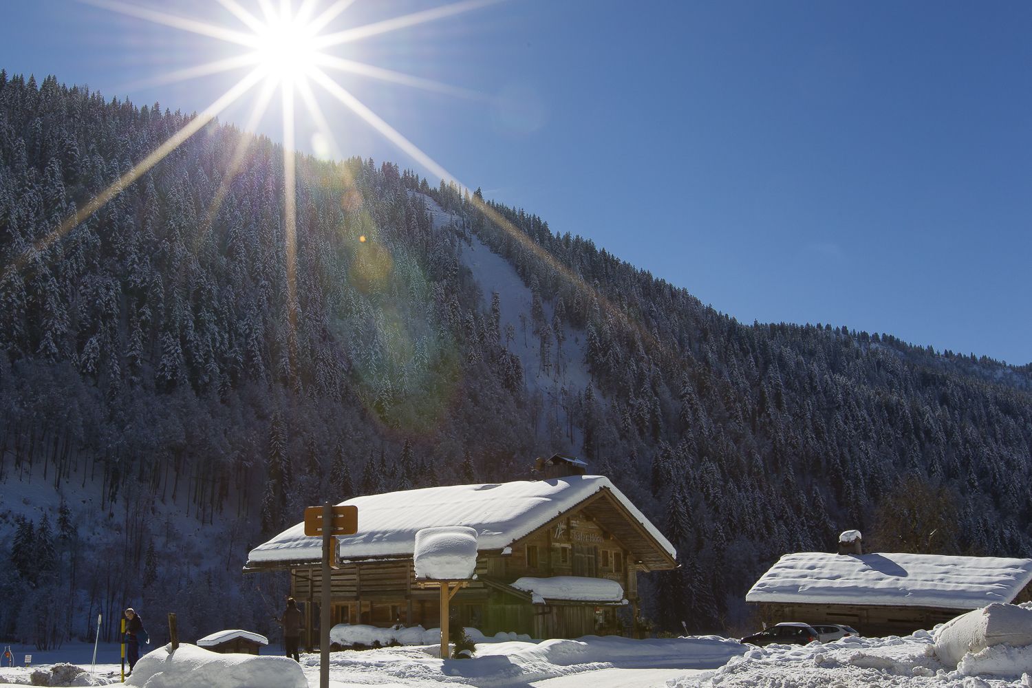 A wooden cabin with a snow-covered roof, surrounded by a snowy landscape and a forested mountain.