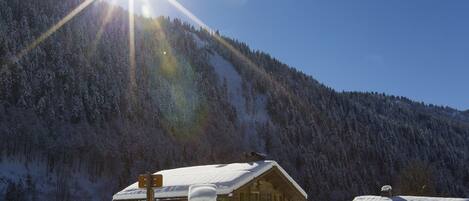 A wooden cabin with a snow-covered roof, surrounded by a snowy landscape and a forested mountain.