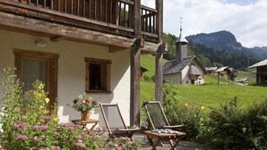 A chalet with a balcony, wooden deck, and a view of a church and mountains. - Les Fermes de Pierre et Anna (Le Grand-Bornand)