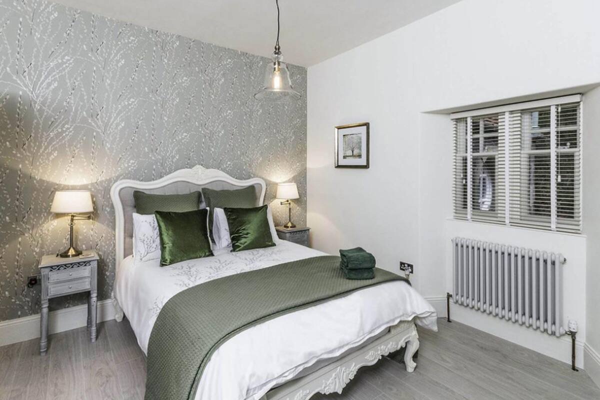 Quarto com cama, duas luminárias de cabeceira, radiador e janela com persianas.