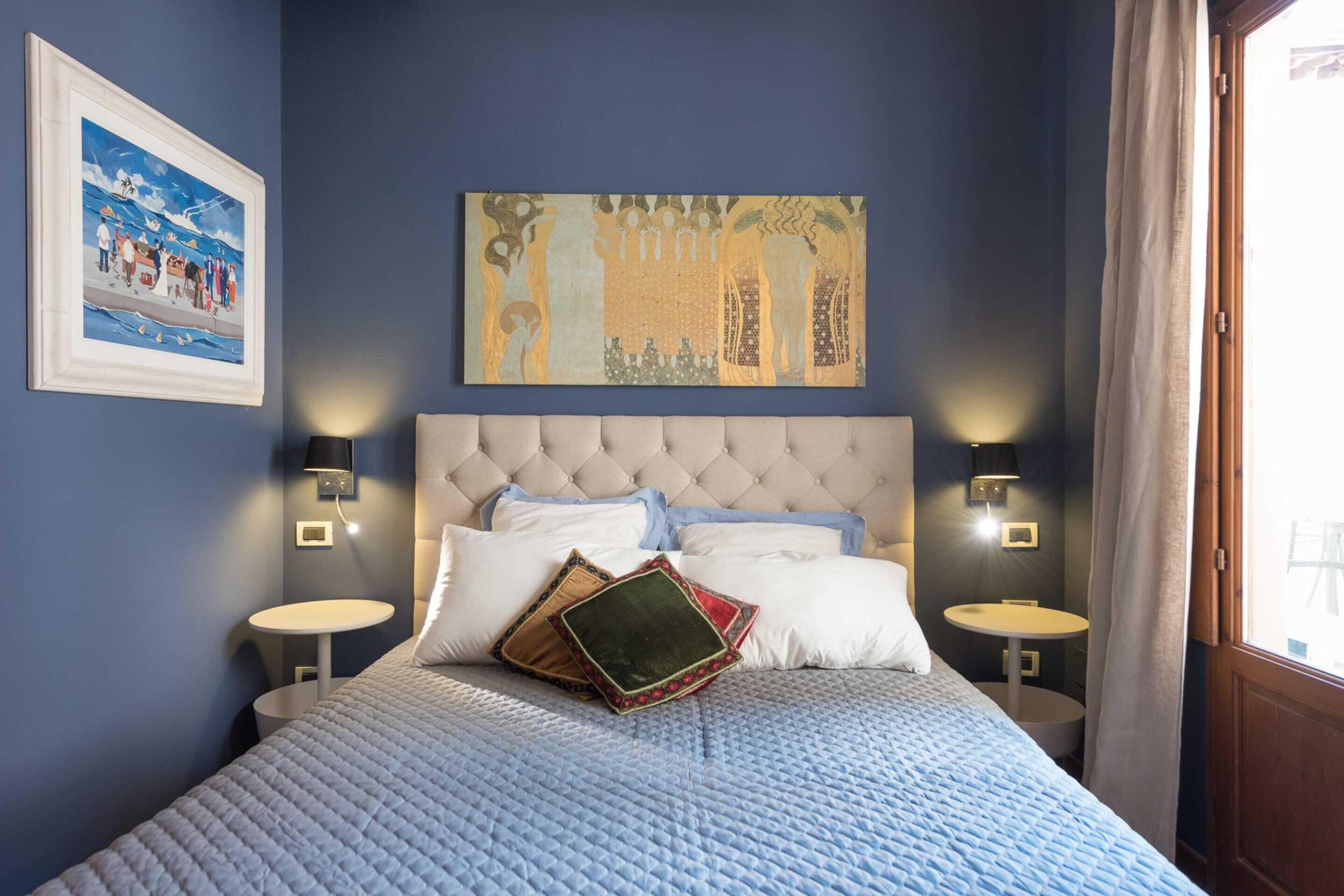 Un dormitorio ordenado con un cabecero capitonado, una cama grande con colcha azul, dos mesitas de noche con lámparas y una obra de arte enmarcada sobre la cama.