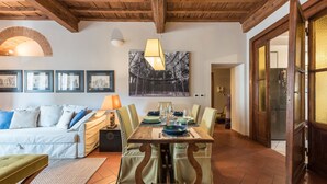 Un comedor con una mesa de madera dispuesta para cuatro personas, rodeada de sillas tapizadas de verde. Un sofá blanco con cojines azules se encuentra en primer plano, y un cuadro enmarcado cuelga de la pared sobre la mesa.