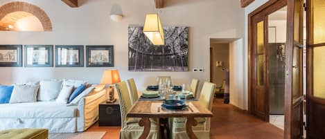 A dining area with a wooden table set for four, surrounded by green upholstered chairs. A white sofa with blue cushions is positioned in the foreground, and a framed picture hangs on the wall above the table.