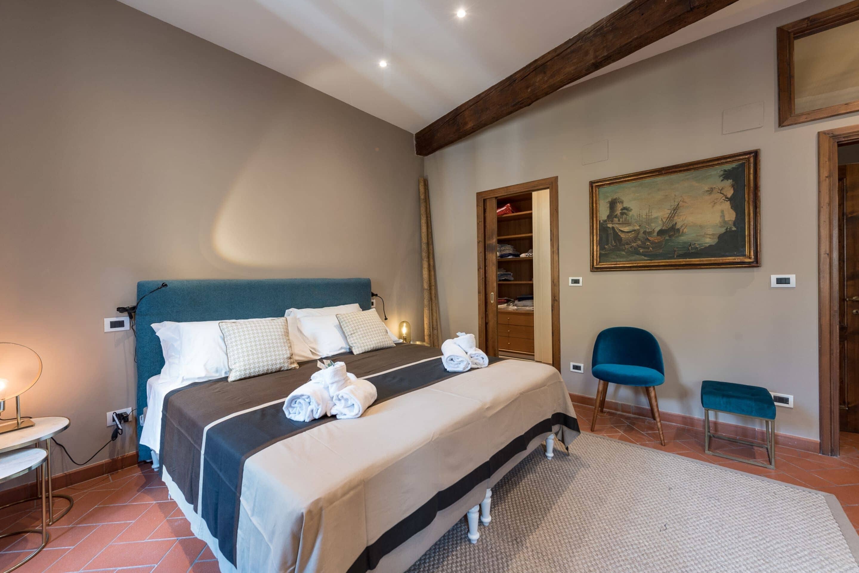 A neatly made bed with a blue headboard, a bedside table with a lamp, a wooden wardrobe, and a painting on the wall.