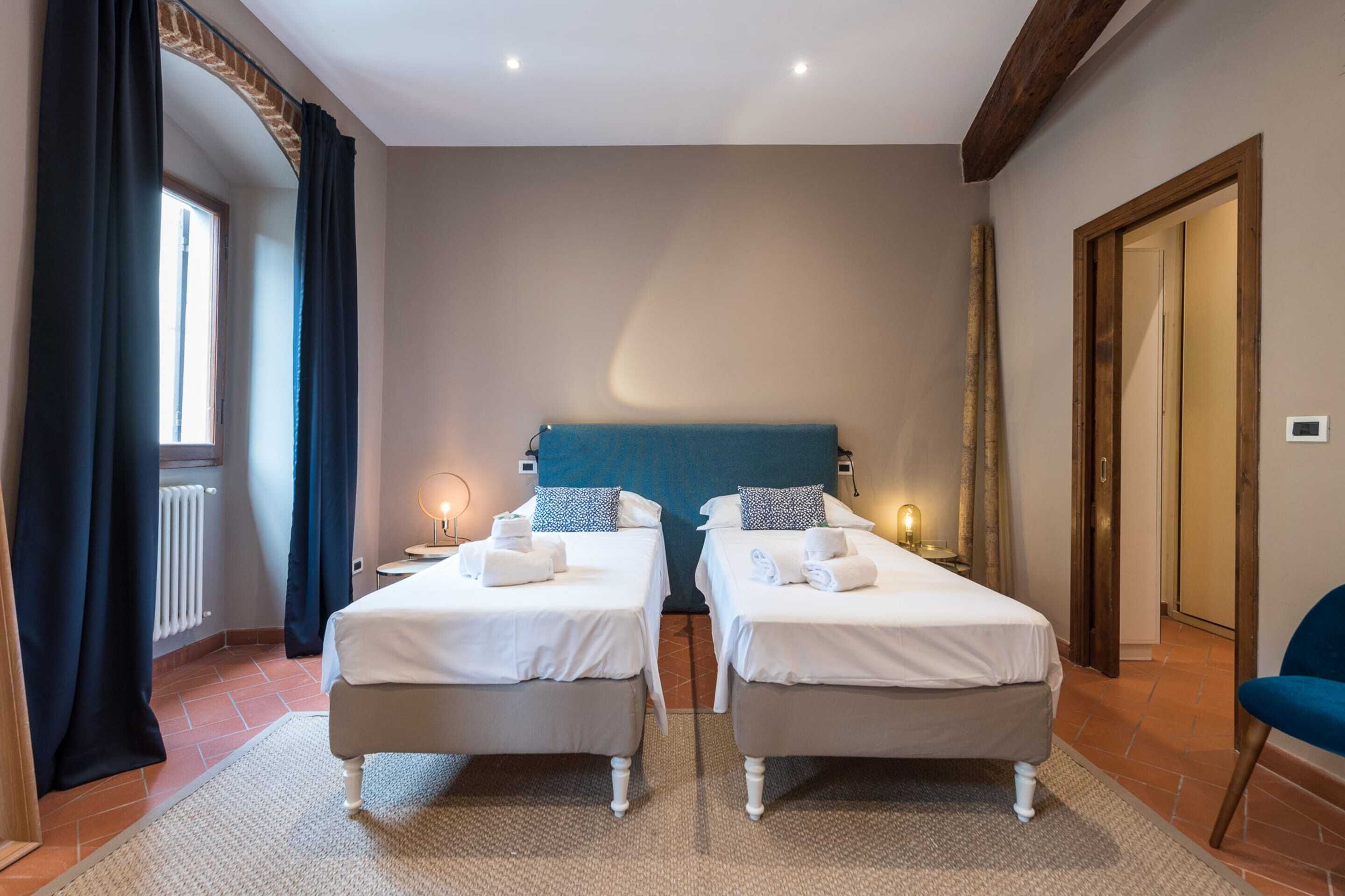 Habitación de hotel con dos camas individuales, un ventanal con cortinas, techo de madera y una silla azul.