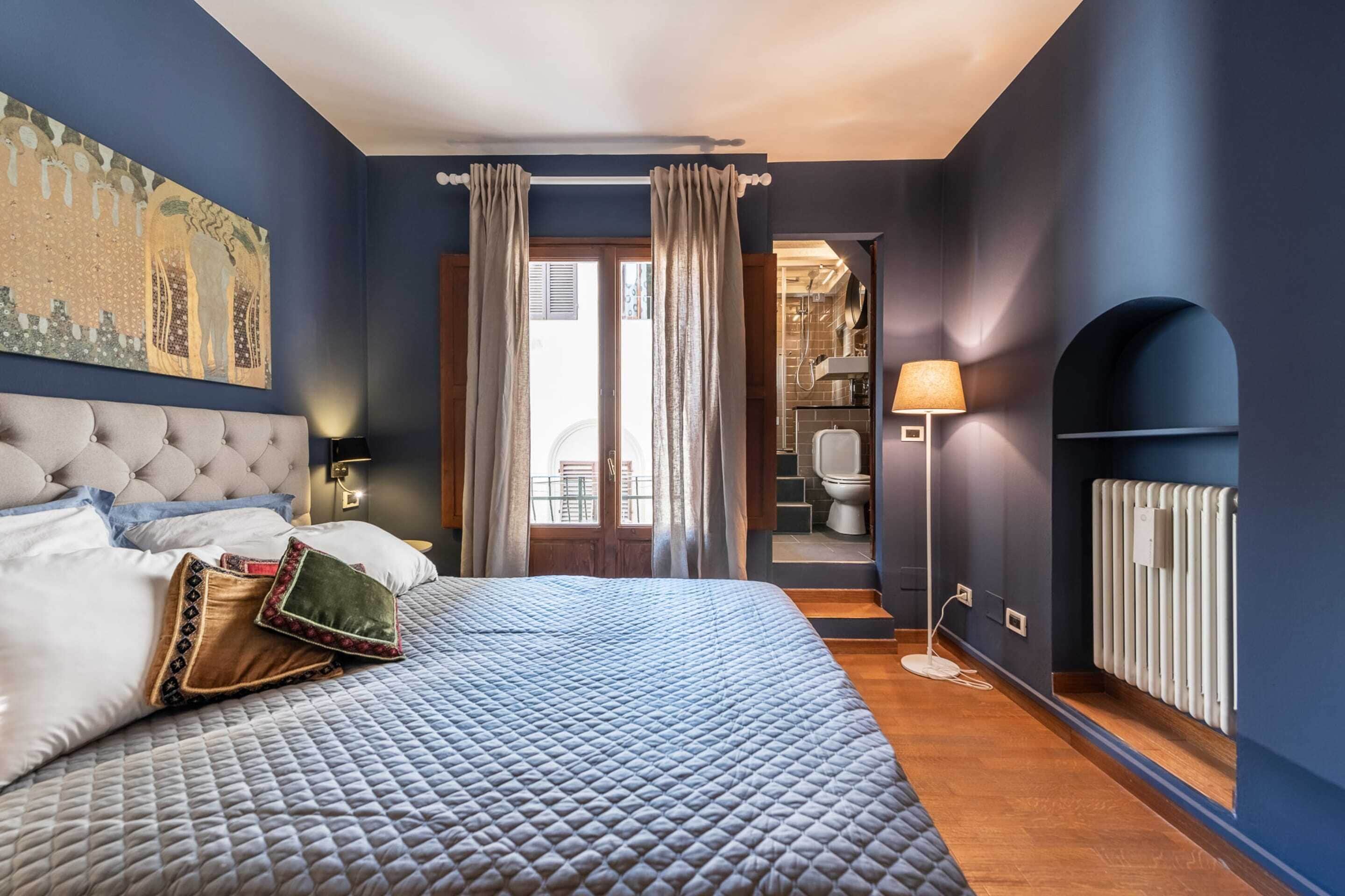 A bedroom with a large bed, a headboard, a nightstand, a lamp, a radiator, and a view of the bathroom.