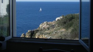 Blick aus einem Zimmer mit einem großen Bogenfenster auf eine Küstenlandschaft, ein Segelboot auf dem Wasser.