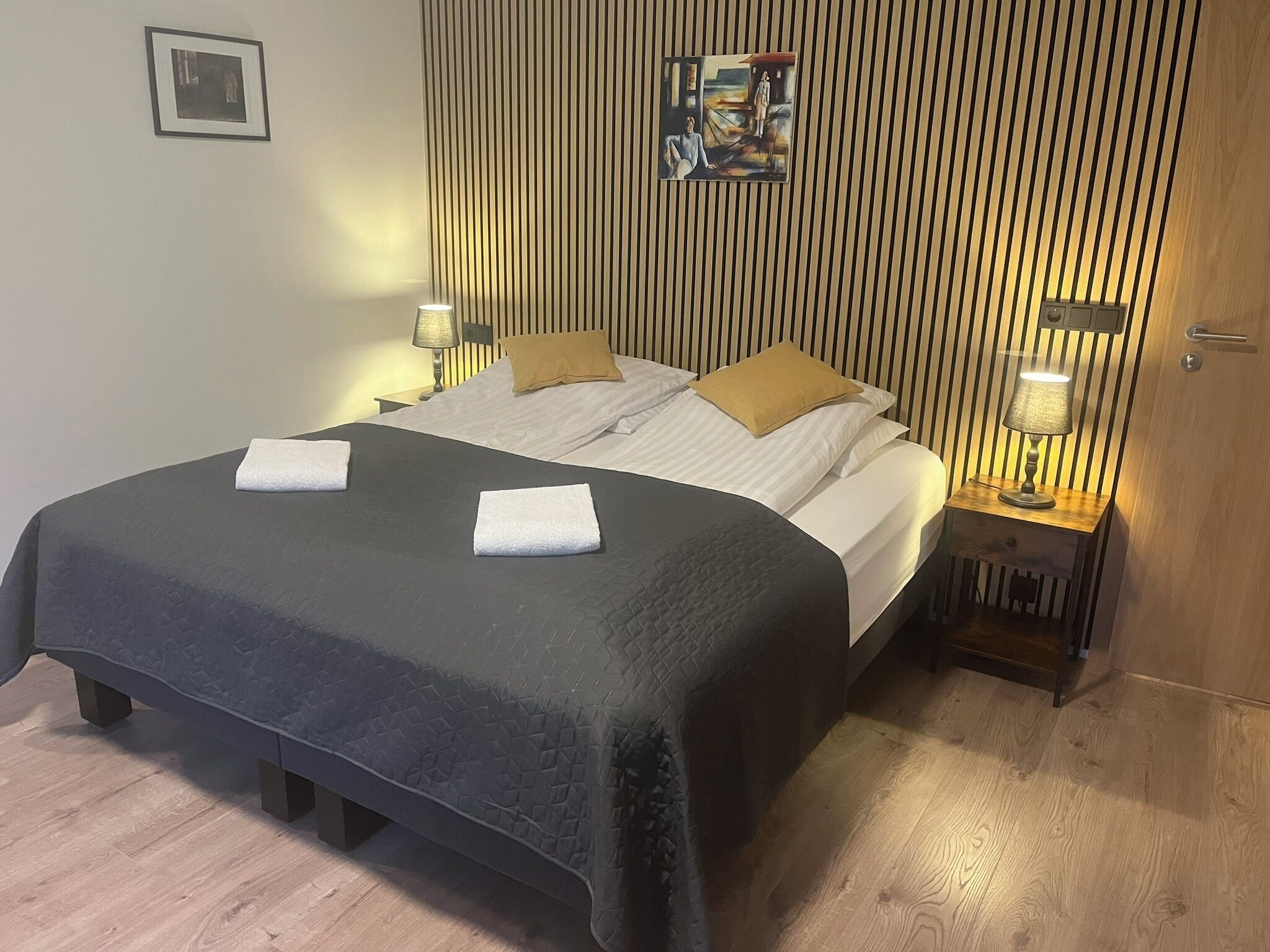 A neatly made bed with a dark quilt, two pillows, and two folded towels. There is a bedside table with a lamp, a framed picture on the wall, and a door with a handle.