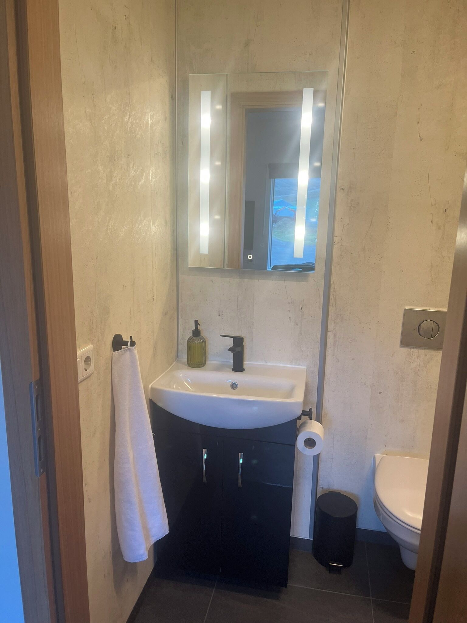 Un baño con un lavamanos blanco, muebles de almacenamiento negros, un espejo, una toalla, un inodoro y un cubo de basura.