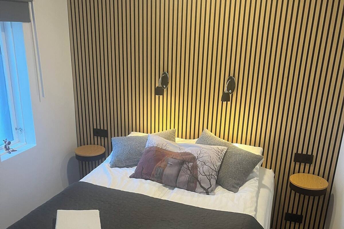 Ein ordentlich gemachtes Bett mit schwarzer Bettdecke und zwei weißen Handtüchern, eine Wand mit vertikalen Holzlatten, zwei Nachttische mit Lampen und ein Fenster links.