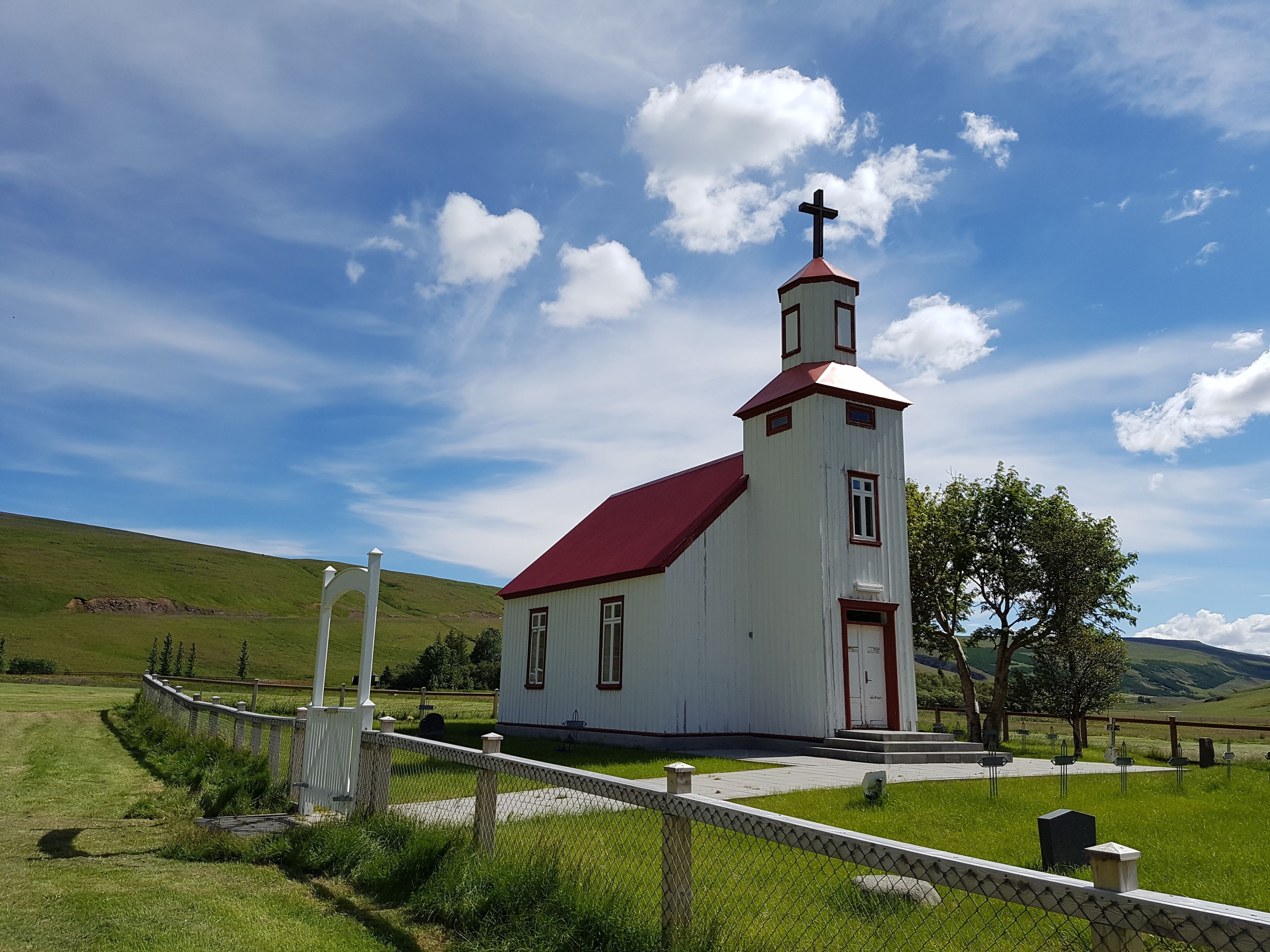 Una iglesia blanca con techo rojo y una cruz en la parte superior, rodeada de colinas verdes y un cielo despejado.