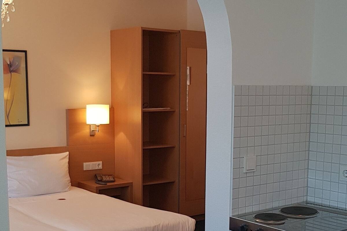 A hotel room with a bed, a desk, a chair, a small table, a lamp, a telephone, a shelf, and a kitchenette.