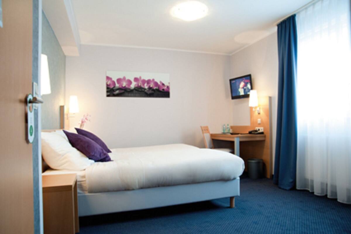A hotel room with a bed, a desk, a chair, a TV, and a painting on the wall.