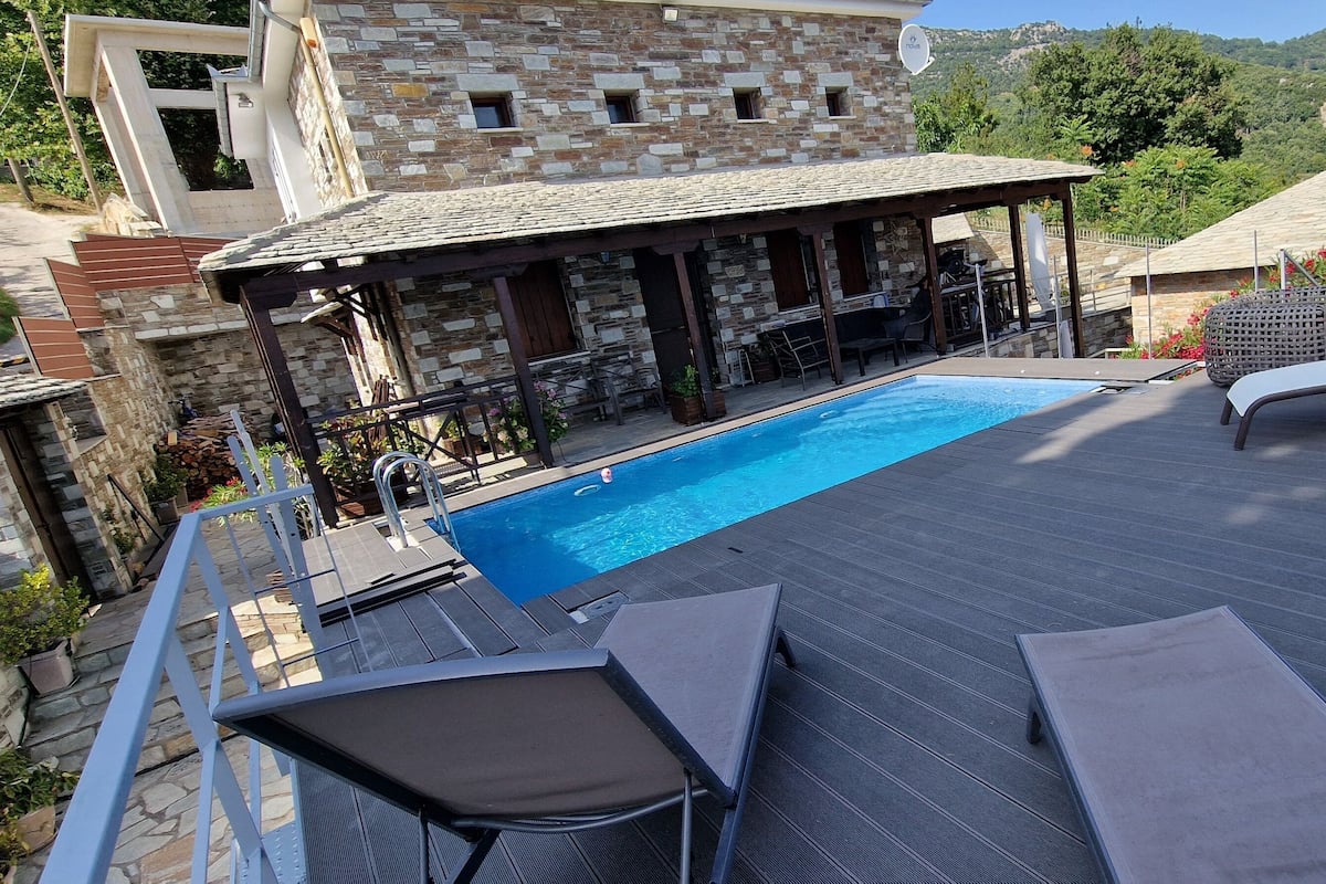 Zona junto a la piscina con sillones de descanso, un espacio de estar cubierto y edificios de piedra al fondo.