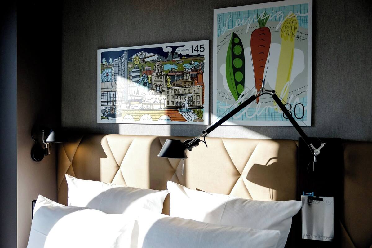 Una habitación de hotel con una cama, un cabecero, una lámpara de pared y dos cuadros enmarcados.