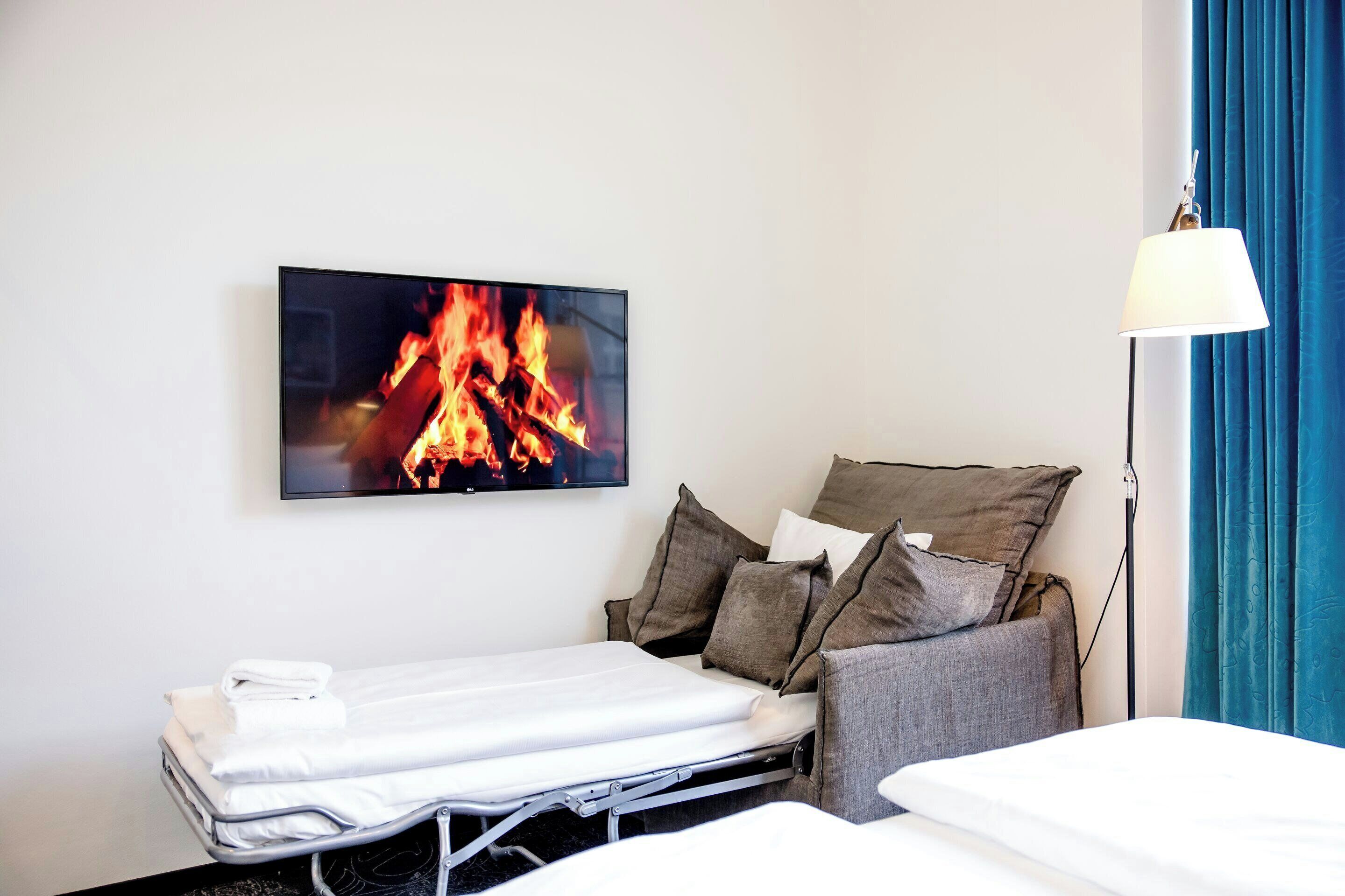 Quarto de hotel com cama, mesa de cabeceira, TV na parede exibindo uma lareira e uma luminária.
