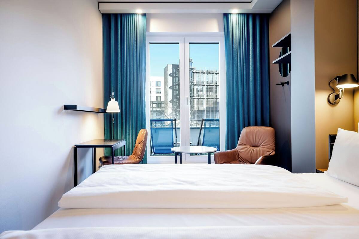 Habitación de hotel con cama, una mesita, dos sillas, ventana con cortinas y vista a edificios.