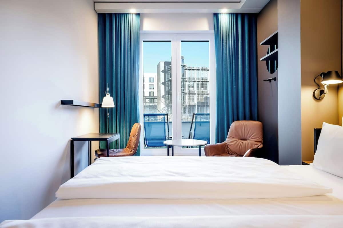 A hotel room with a bed, a small table, two chairs, a window with curtains, and a view of buildings.