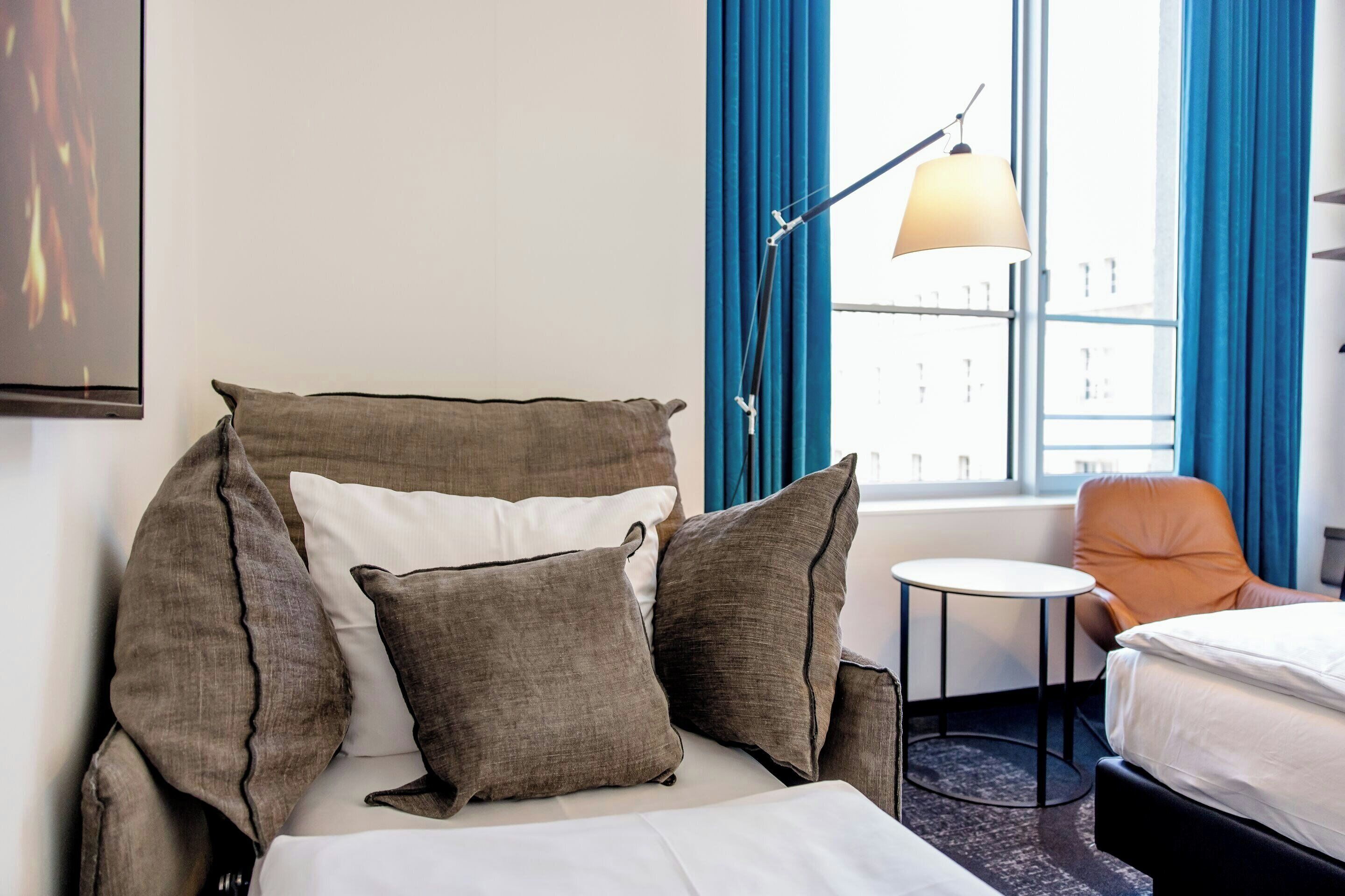A hotel room with a bed, pillows, a small table, a chair, a lamp, and a window with curtains.