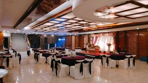A spacious banquet hall with multiple round tables, each with a white tablecloth and black sashes. The room features a high ceiling with recessed lighting and a stage area with floral decorations. - Swasno By Rivlet (Gurugram)
