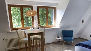A modern loft bedroom with a sloped ceiling, a dining area with a table and chairs, a blue armchair, and a large window with a view of trees.