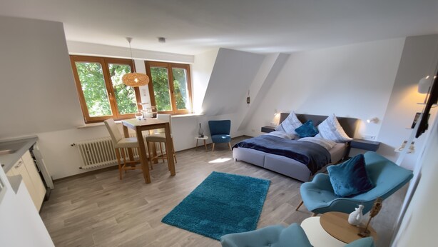 A modern loft apartment with a dining area, a bedroom with a large bed and blue accent pillows, and a kitchen with a white countertop and wooden cabinets.