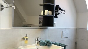 A modern bathroom with a white sink, a mirror, a black shelf with toiletries, a toilet, and a small trash bin.