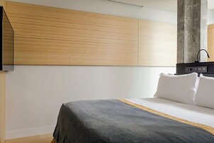 Quarto moderno com uma cama, painel de parede de madeira e um dispositivo eletrônico preto sobre um suporte.