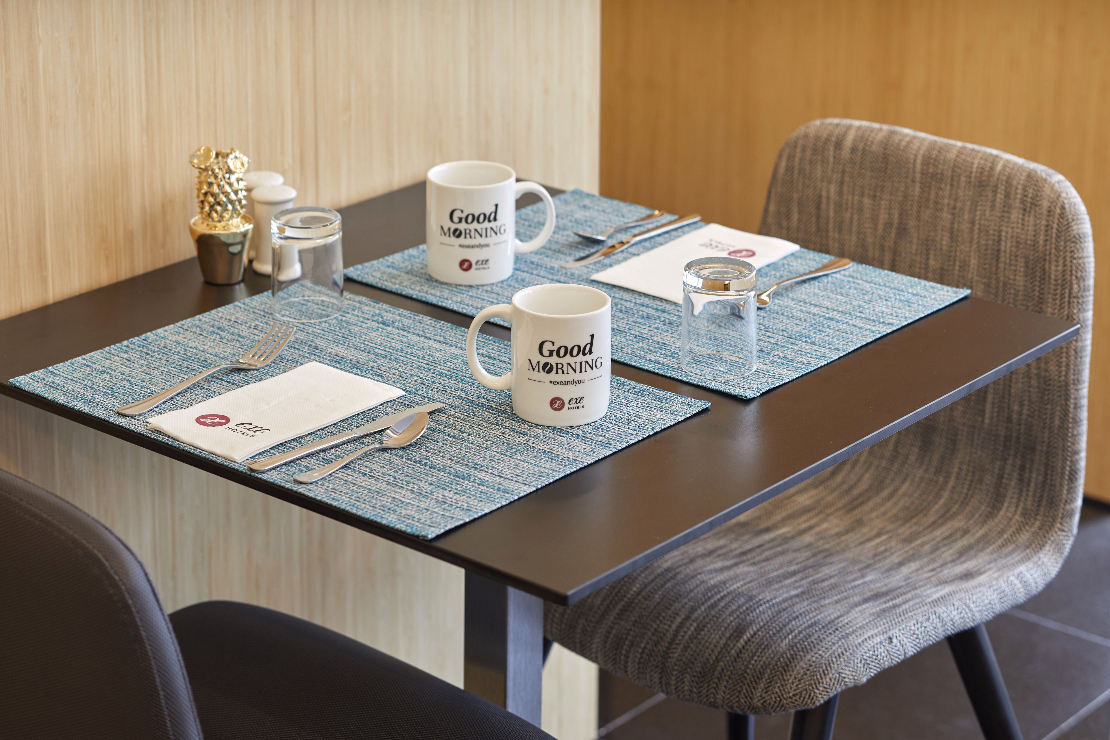 A neatly set dining table with two white mugs, a fork, a knife, and a glass.
