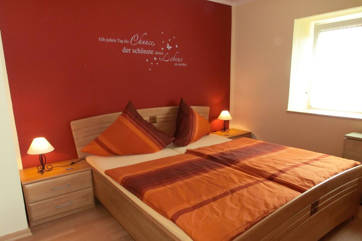 Cama bem arrumada com manta listrada, duas luminárias de cabeceira e parede com frase motivacional.
