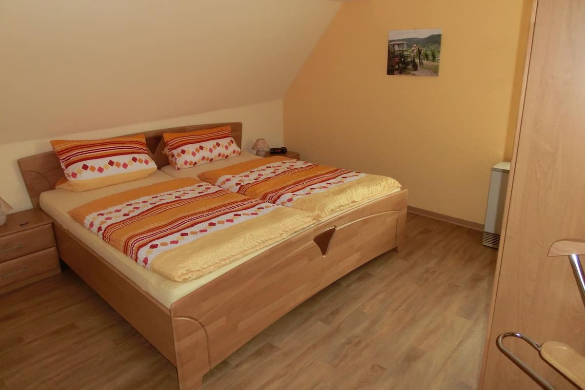 Quarto com cama de madeira, duas almofadas, mesa de cabeceira e um pequeno criado-mudo.