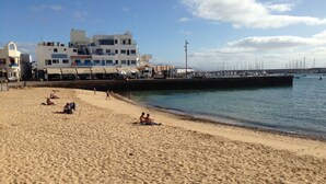 A sandy beach with people sunbathing and walking. There are buildings in the background and a pier with boats.
