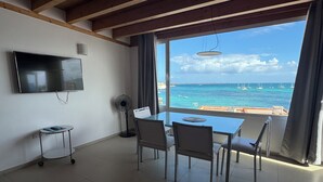 A modern room with a large window overlooking the sea, a flat-screen TV mounted on the wall, and a dining table with chairs.