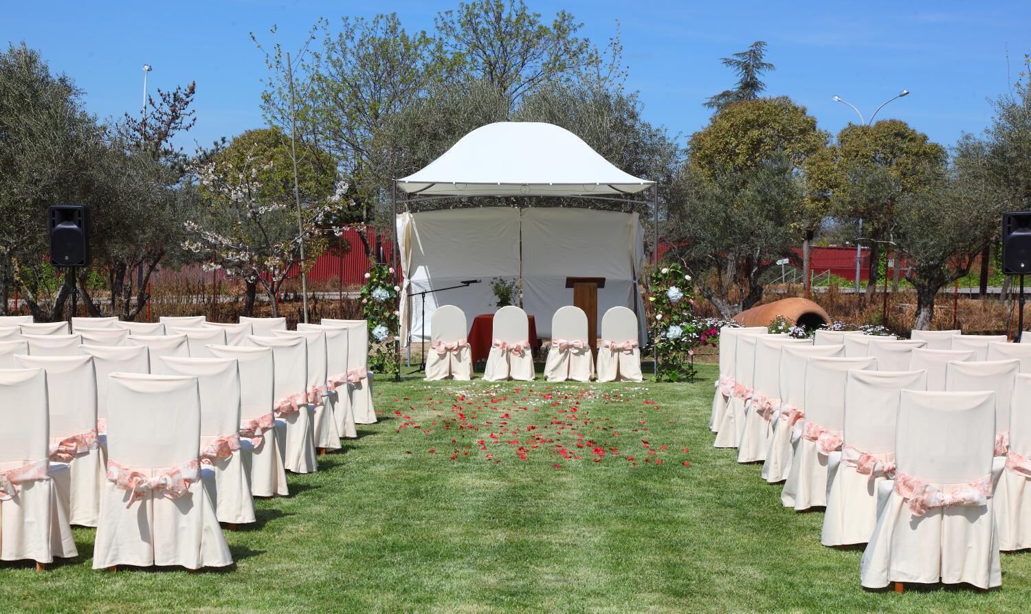 Una carpa con dosel blanco y una puerta pequeña, rodeada de hileras de sillas blancas con cintas rosas, sobre una zona cubierta de césped con árboles al fondo.