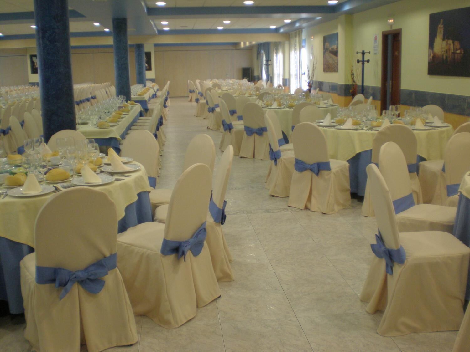 A banquet hall with rows of tables set for a formal event, each with white tablecloths and blue sashes on chairs.