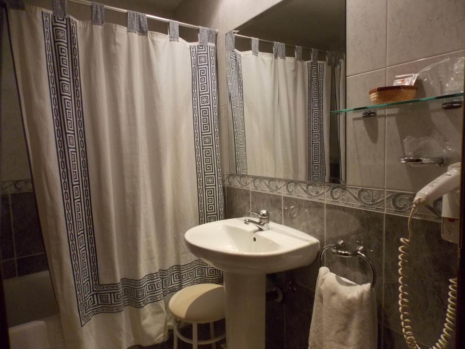 Un baño con un lavabo blanco, un espejo, una cortina de ducha con un motivo geométrico, un estante de vidrio con artículos de aseo y un toallero.