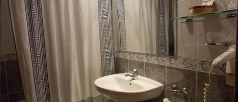 Un baño con un lavabo blanco, un espejo, una cortina de ducha con un motivo geométrico, un estante de vidrio con artículos de aseo y un toallero.