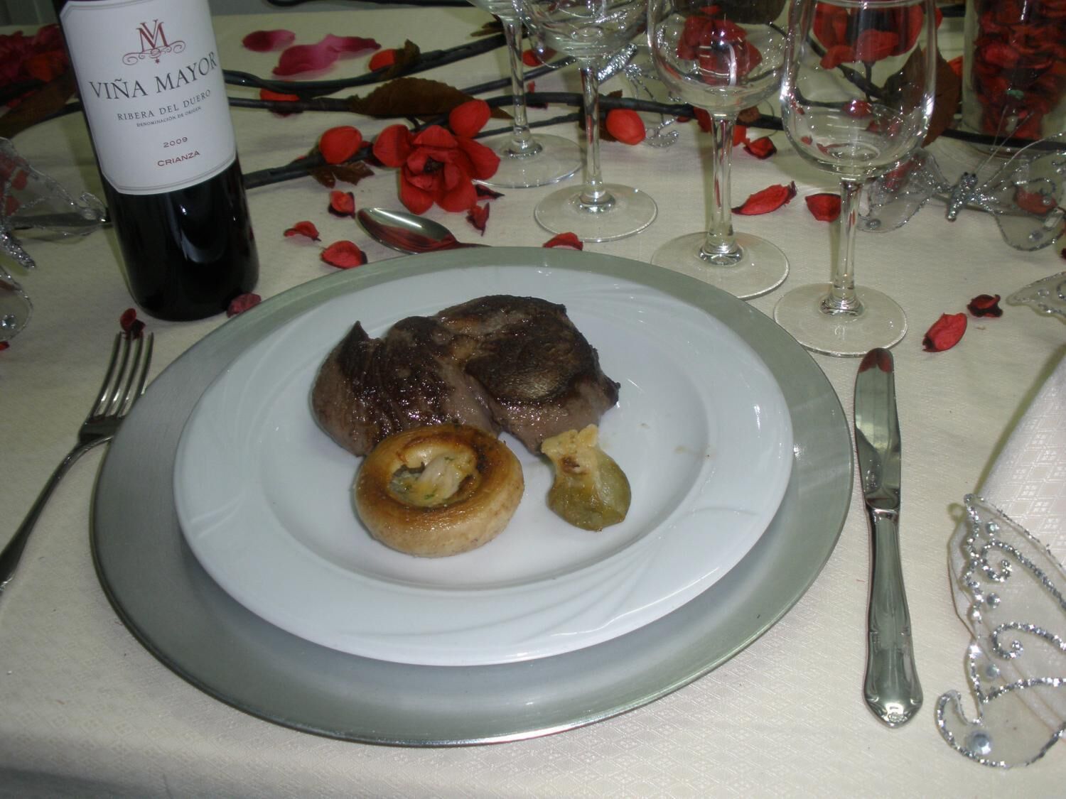 A plate with steak and a small baked item, a bottle of wine, and wine glasses on a table with rose petals.